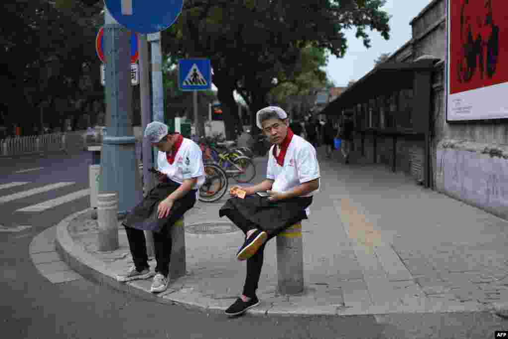 2018年8月22日，北京，两名餐馆工作人员坐在路边的石墩上。他们身后是关于马克思和恩格斯的宣传画。他们所在的餐馆如果是民营的而且雇佣职工至少8人，按照马克思的理论，就是剥削。马克思这一论点在改革开放初期曾经阻碍了中国私营企业的发展，后来被突破了，一大原因是要解决就业问题。